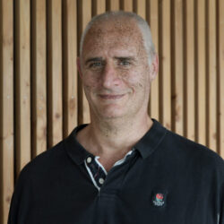 Représente un homme en polo noir, souriant devant un mur de bois - Agrandir l'image, fenêtre modale